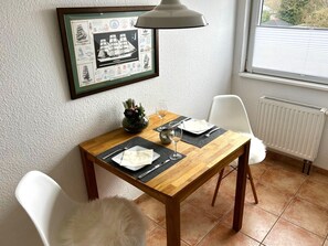 Un comedor con una mesa de madera dispuesta para dos comensales, un mapa náutico enmarcado y una ventana con persianas.