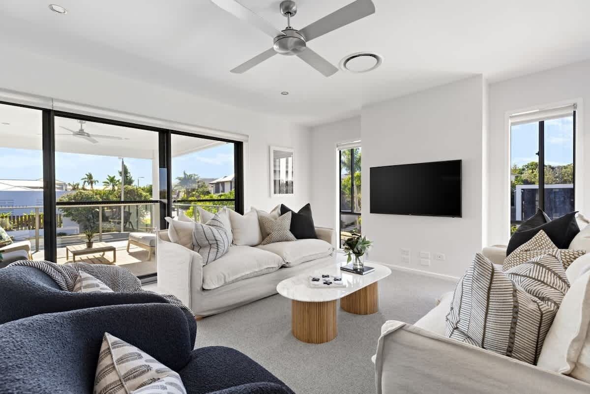 A modern living room with a sofa, armchair, and coffee table, a flat-screen TV mounted on the wall, and a ceiling fan.