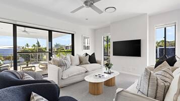 A modern living room with a sofa, armchair, and coffee table, a flat-screen TV mounted on the wall, and a ceiling fan.