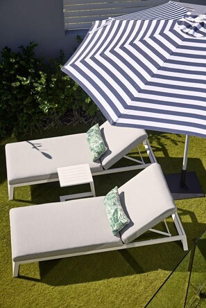 Two white lounge chairs with green patterned cushions, an umbrella with a chevron pattern, and a backdrop of green foliage. - Sirius (Kingscliff)