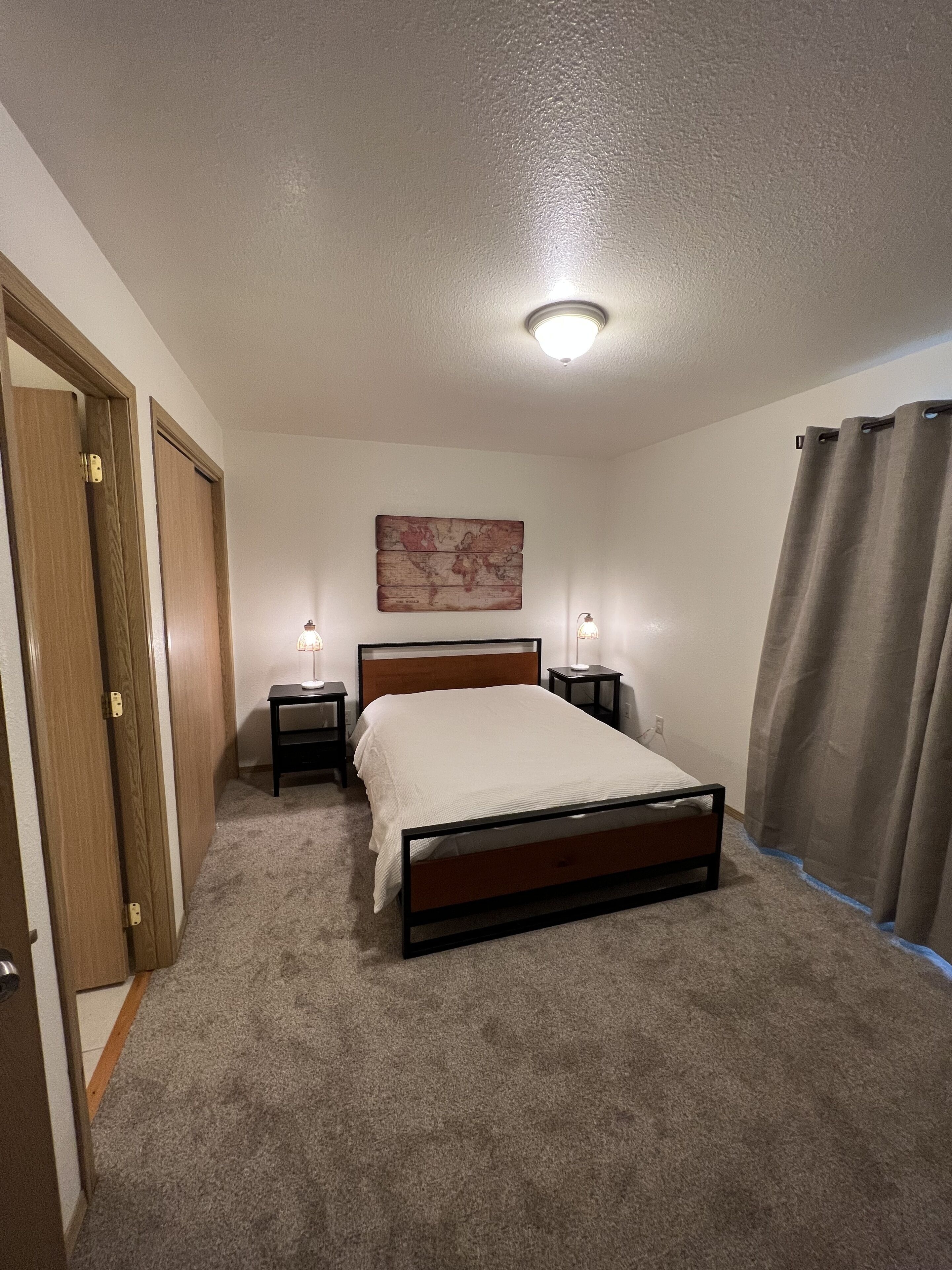 A bedroom with a bed, two bedside tables, a nightstand, a wall-mounted light, a ceiling light, and a door.