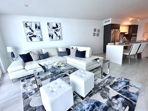 Ein modernes Wohnzimmer mit einem weißen Ecksofa, einem Glastisch und Poufs. An den Wänden hängen abstrakte Gemälde, und im Hintergrund ist eine Küchenzeile zu sehen.