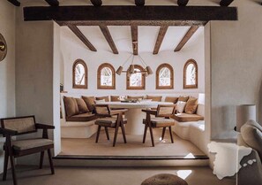 A cozy interior with a round table surrounded by cushioned seating, wooden beams on the ceiling, and arched windows.