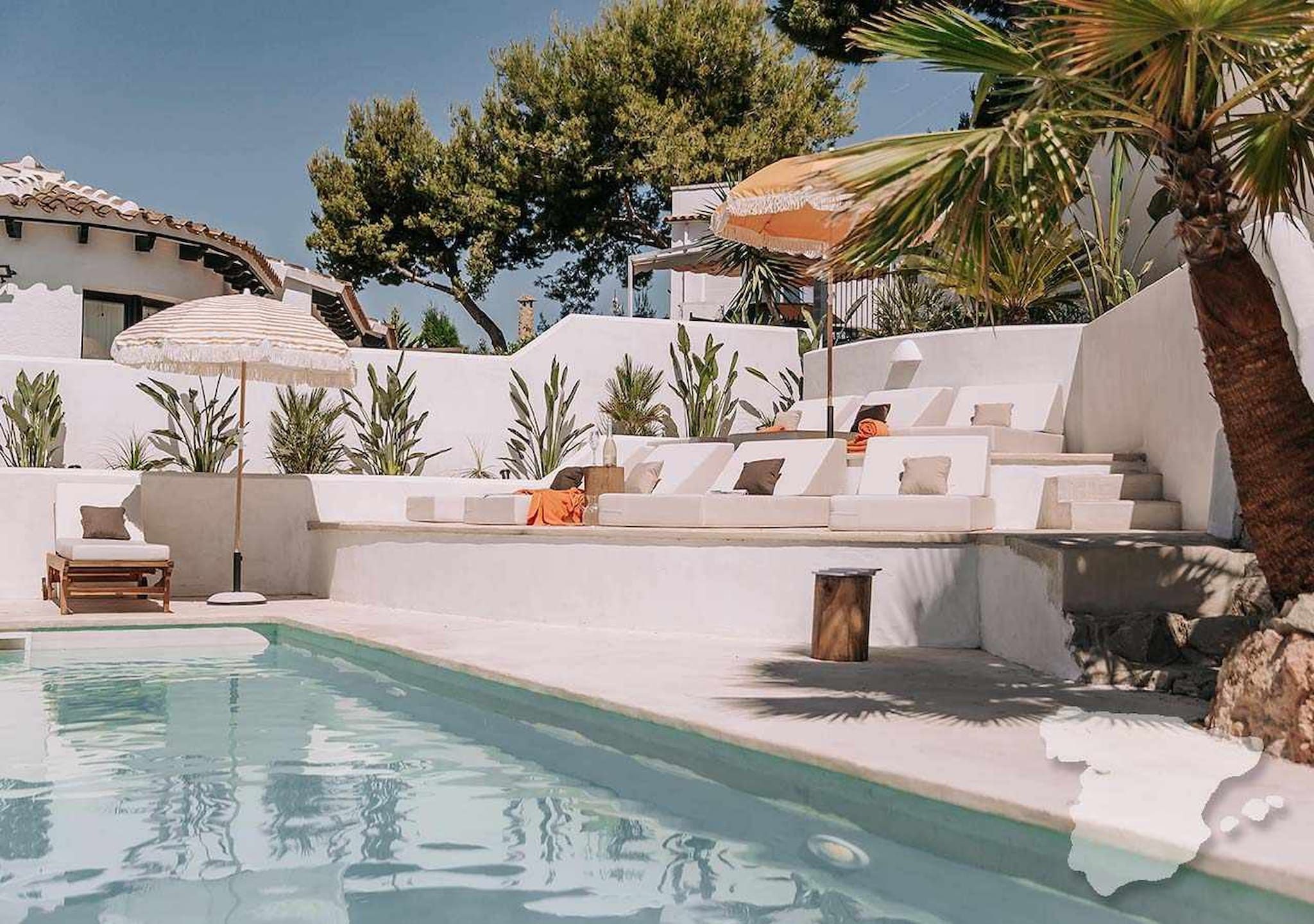 A pool area with lounge chairs and a small table, surrounded by white walls and plants.