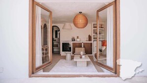 Una habitación con un amplio marco de puerta de madera que da a otro espacio, una lámpara colgante de fibras tejidas, una pequeña mesa con un jarrón y una alfombra blanca.