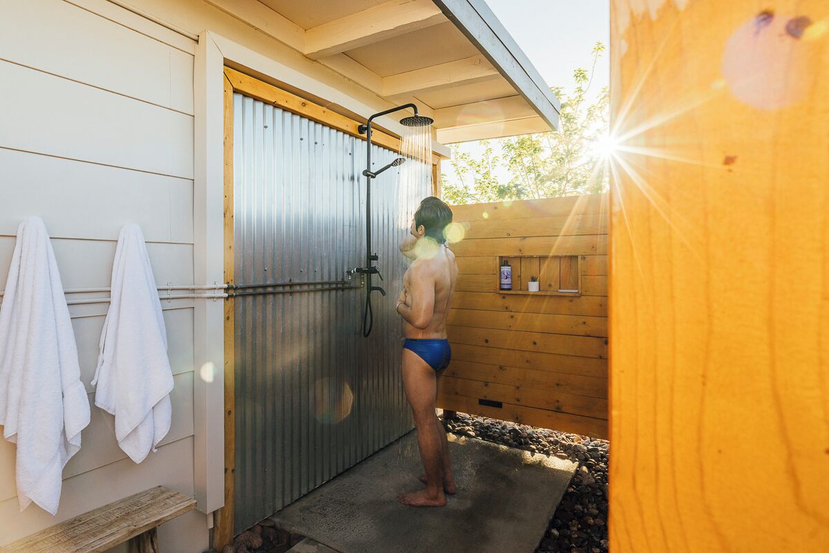 A person in swim trunks standing under a shower in a wooden cabin with towels hanging on the wall.