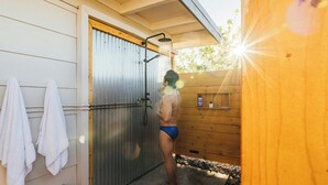 A person in swim trunks standing under a shower in a wooden cabin with towels hanging on the wall.