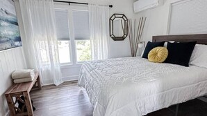 Un dormitorio con cama, ventilador de techo, ventana con cortinas, mesita de noche y suelo de madera.