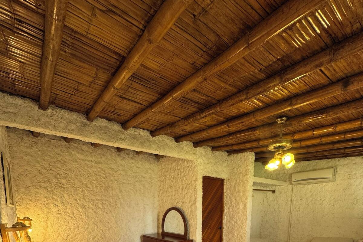 A room with two beds, a wooden ceiling, a white textured wall, and a small wooden nightstand with a lamp.
