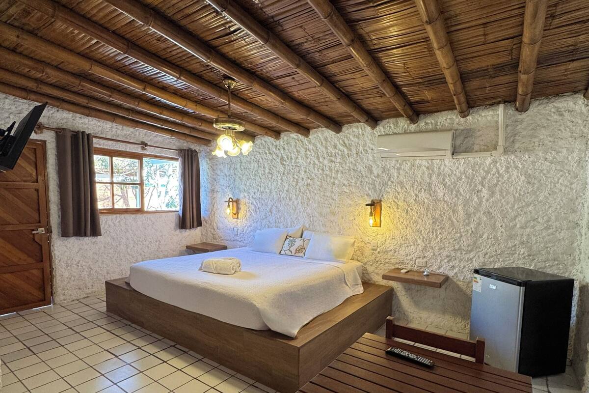 A bedroom with a bed, a wooden table, a small fridge, and a window.