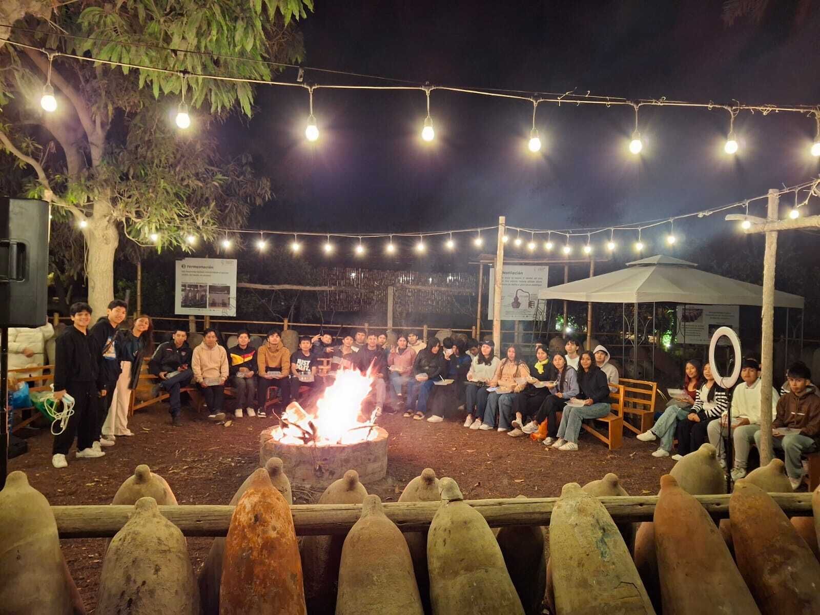 An outdoor gathering with a fire pit, people seated on benches, and string lights overhead.