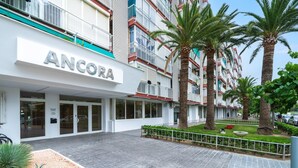 A modern building with the name "ANCORA" prominently displayed. It features a spacious entrance with glass doors, a paved walkway, and a small garden area with palm trees and shrubs. - Romantic loft sea views 8km from Port Aventura (La Pineda)