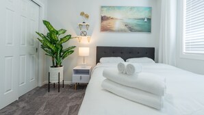A neatly arranged bedroom with a bed, a nightstand, a lamp, a potted plant, and a painting of a beach scene.