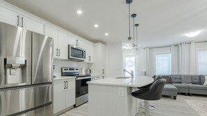 A modern kitchen with stainless steel appliances, a central island with bar stools, and a grey sofa in the adjacent living area.