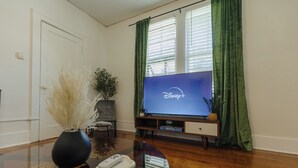 Een woonkamer met een flatscreen televisie waarop het Disney+-logo staat, een vaas met gedroogde planten, een afstandsbediening en een houten salontafel.