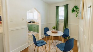 Een eetgedeelte met een ronde tafel, vier blauwe stoelen, een kleine plant in een pot en een groen gordijn.