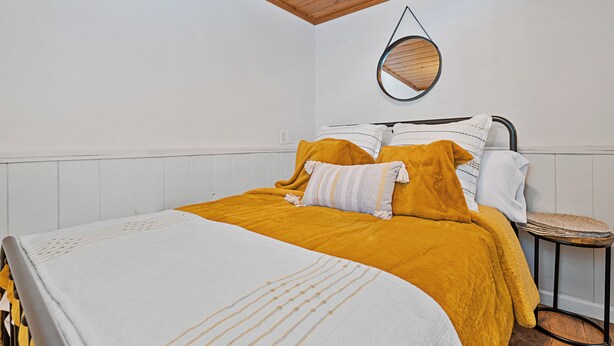 A neatly made bed with a white and yellow bedspread, a round mirror on the wall, and a small stool beside the bed. - The Vintage Bear Cabin (Big Bear City)