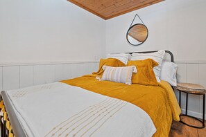 Een keurig opgemaakt bed met een wit en geel sprei, een ronde spiegel aan de muur en een klein krukje naast het bed.