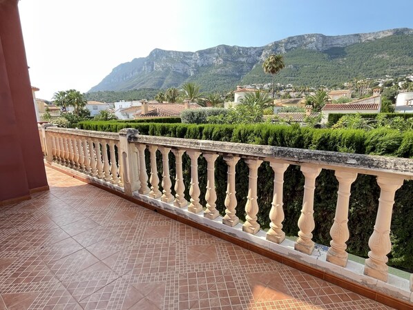 Un balcón con suelo de baldosas y barandilla, con vista a un paisaje montañoso con casas y vegetación.