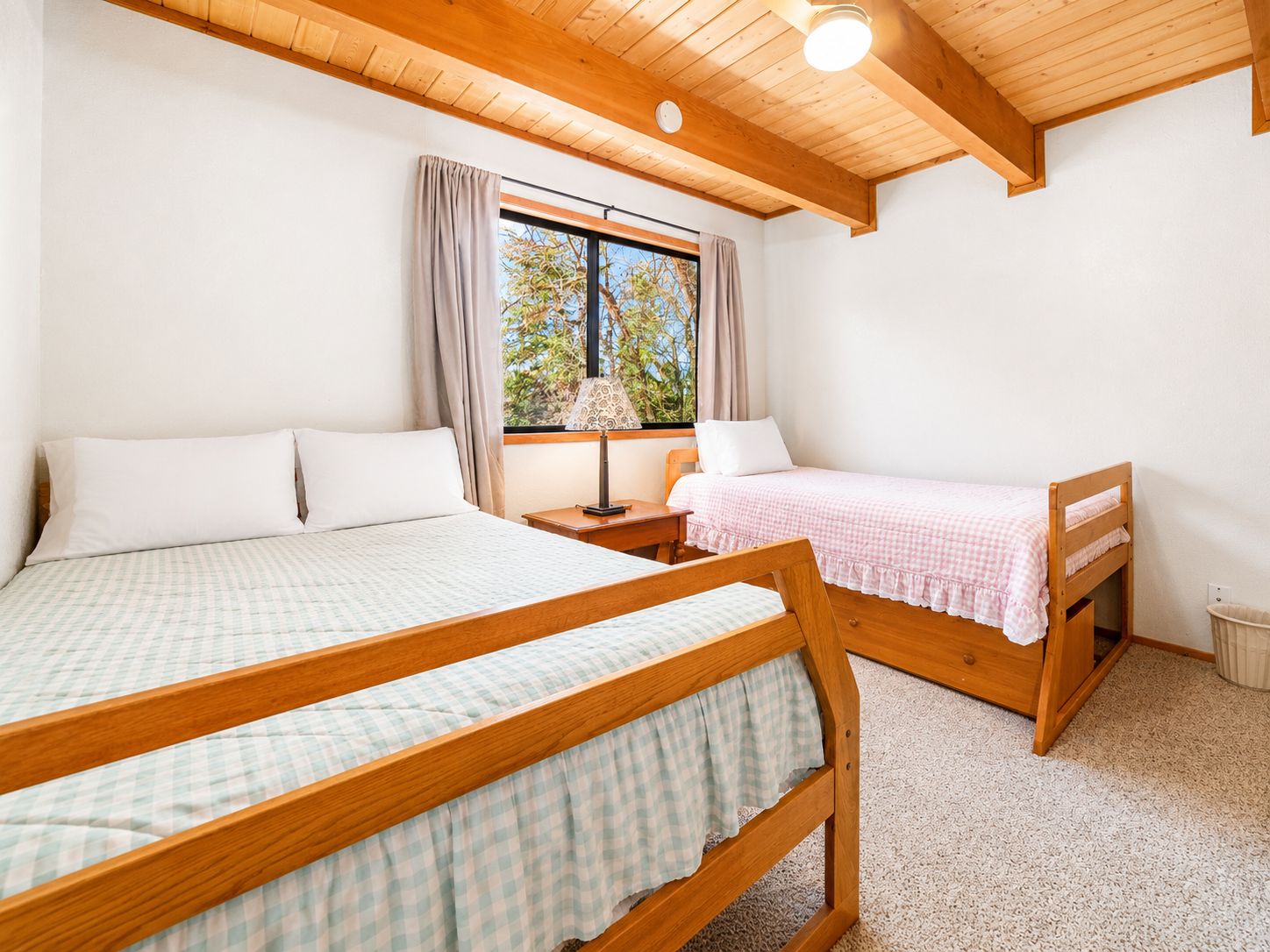 A bedroom with two wooden beds, a window with curtains, a nightstand, and a lamp.