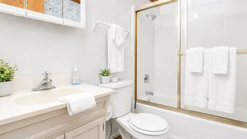 Baño con lavamanos blanco, inodoro y bañera. Cuenta con una cabina de ducha con mampara de vidrio y marco dorado.