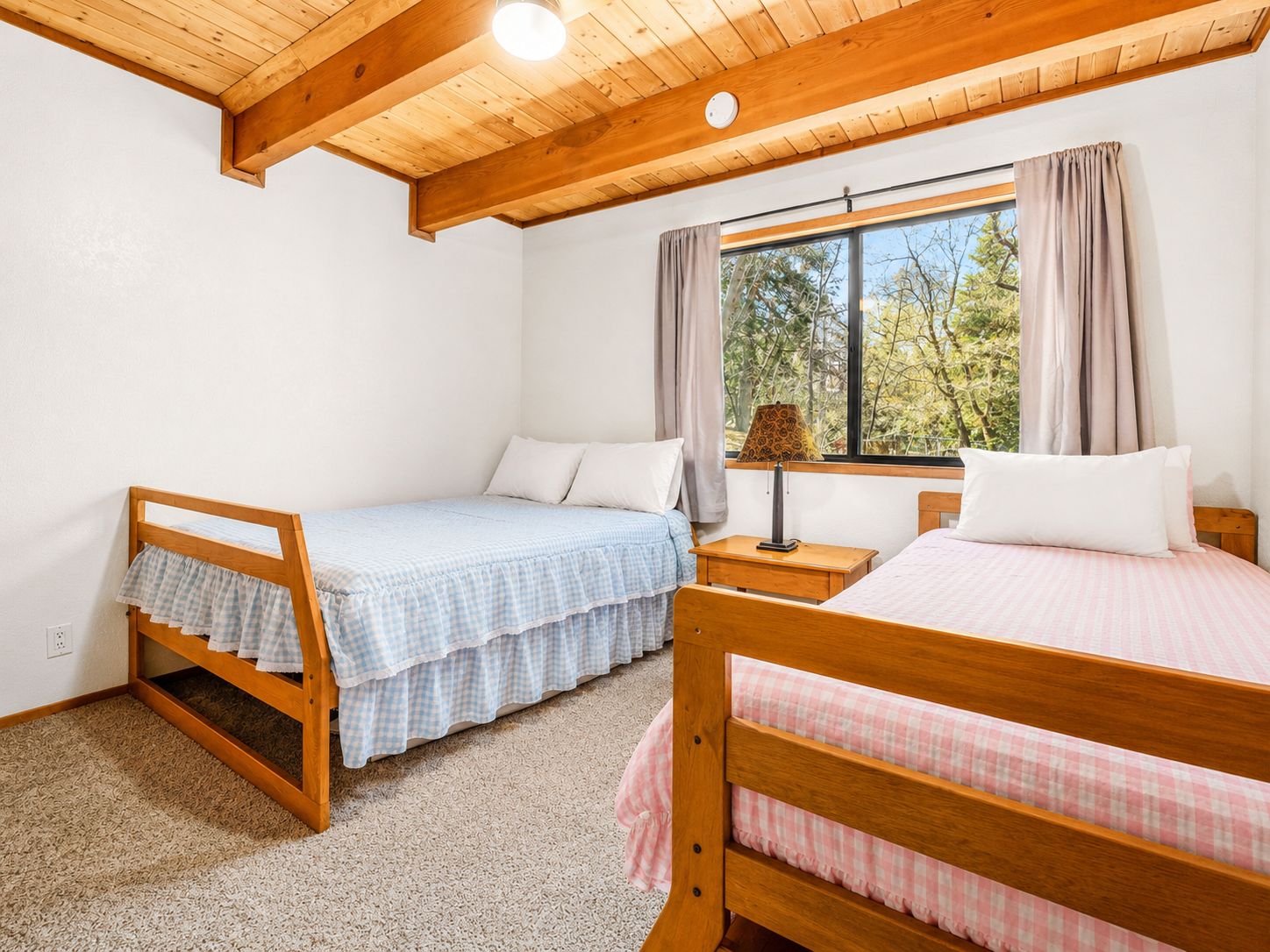 A bedroom with two wooden beds, a window with curtains, and a lamp on a bedside table.