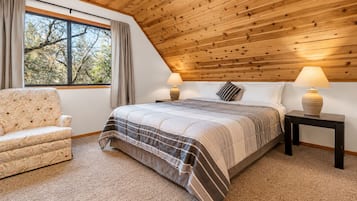 Un dormitorio con techo inclinado, una cama con ropa de cama a rayas, una silla, dos mesitas de noche con lámparas y una ventana con cortinas.