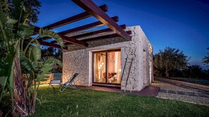A modern house with a stone exterior, a glass door leading to a room with a bed and a chair, and a pergola with wooden beams. - Villa Sumac 6 Emma Villas (Sortino)