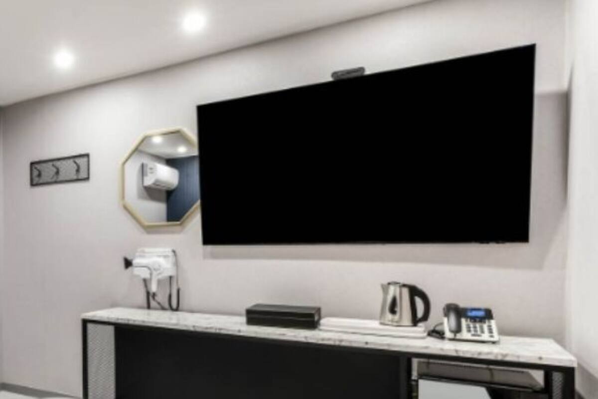 A hotel room with a door, a small table, a chair, a vanity with a mirror, a hairdryer, a kettle, and a flat-screen TV mounted on the wall.