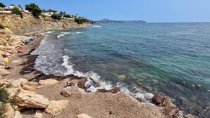 A coastal beach with rocky terrain, clear blue water, and a distant mountain range.