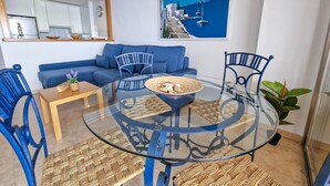 A modern living room with a glass-top dining table, blue chairs, a sofa, and a painting of a coastal scene.