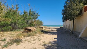 Un sentier sablonneux mène à une plage, avec des piles de planches de bois et un bâtiment doté d’un toit en pente sur la droite.