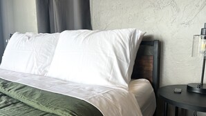 Ein ordentlich bezogenes Bett mit weißen Leinentüchern und einer grünen Tagesdecke, ein Nachttisch mit einer Lampe sowie eine strukturierte Wand.