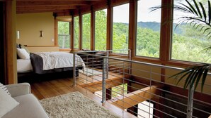 Un dormitorio con cama, vistas al bosque a través de amplias ventanas y un balcón con barandilla.