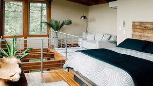 Un dormitorio con techo de madera, una cama con ropa de cama estampada, una mesita de noche, un escritorio con una planta y una escalera con barandilla.