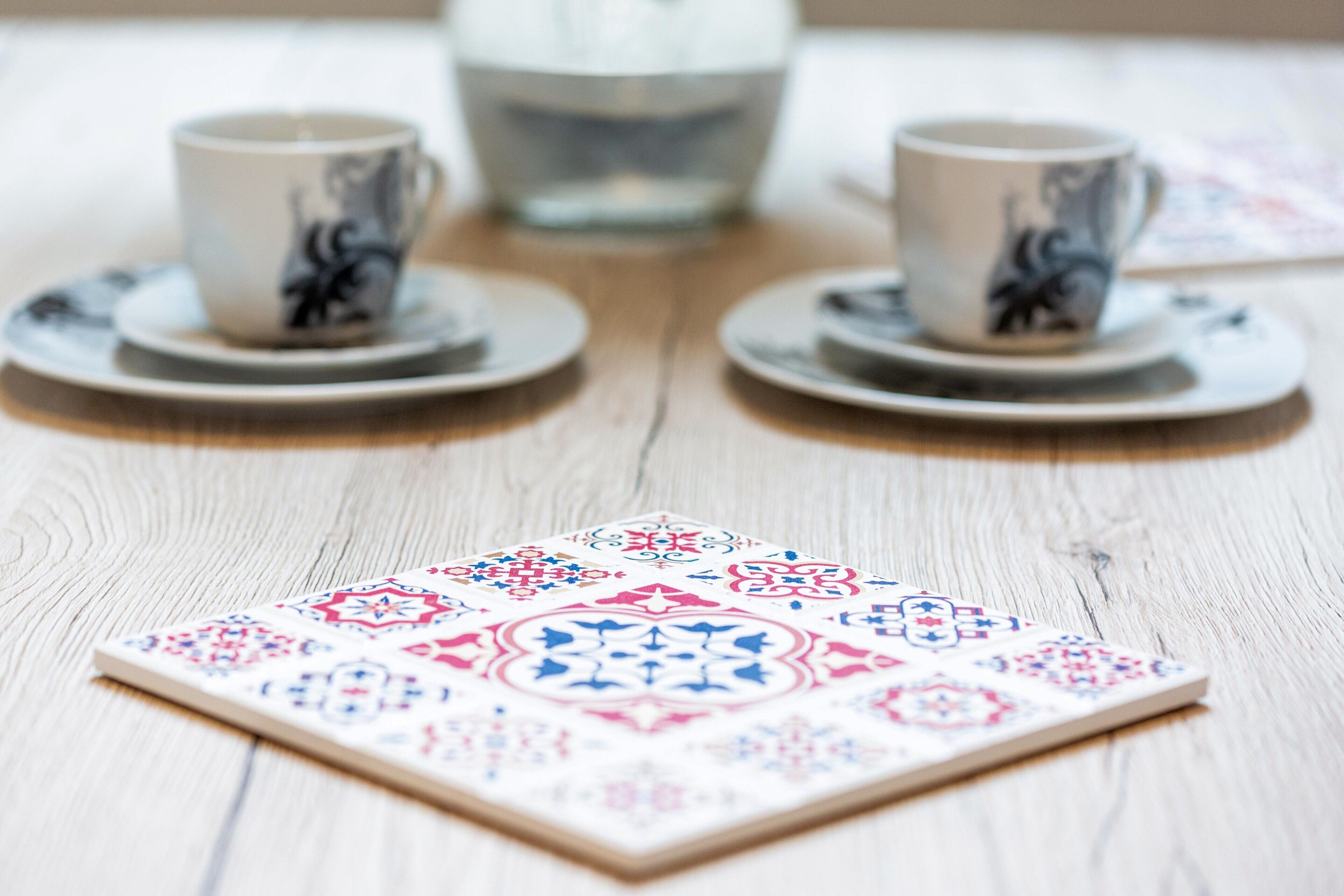 A tabletop with a patterned coaster, two cups with saucers, and a glass.