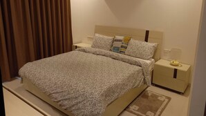 A neatly made bed with a patterned bedspread, a bedside table with a lamp, and a wall-mounted light fixture. - 1Bed Apartment - Kalu Beach Resort, Parking (Salalah)