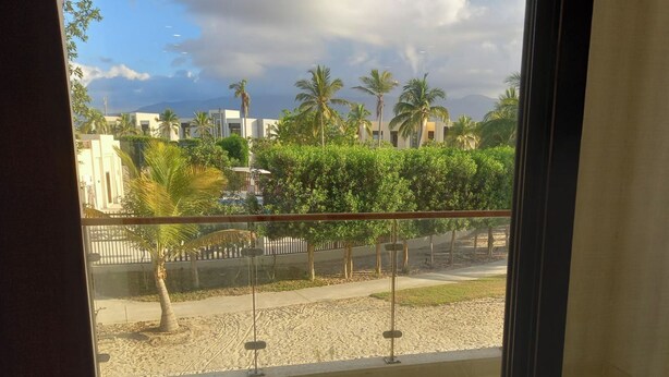 A view from a window overlooking a sandy area with palm trees and a building in the background. - 1Bed Apartment - Kalu Beach Resort, Parking (Salalah)
