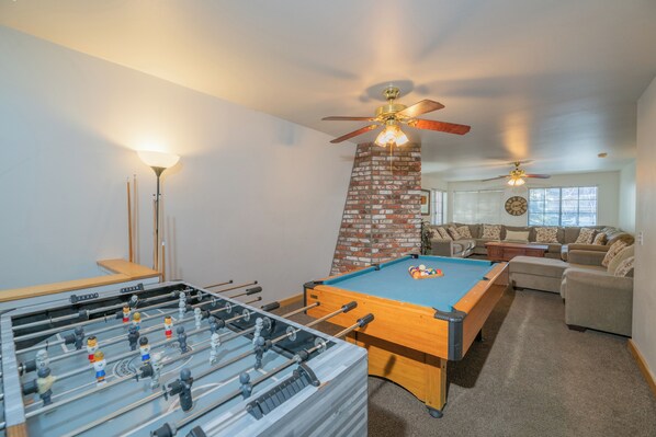 Une pièce équipée d’un baby-foot, d’une table de billard, d’une cheminée en briques, d’un ventilateur de plafond et d’un canapé.