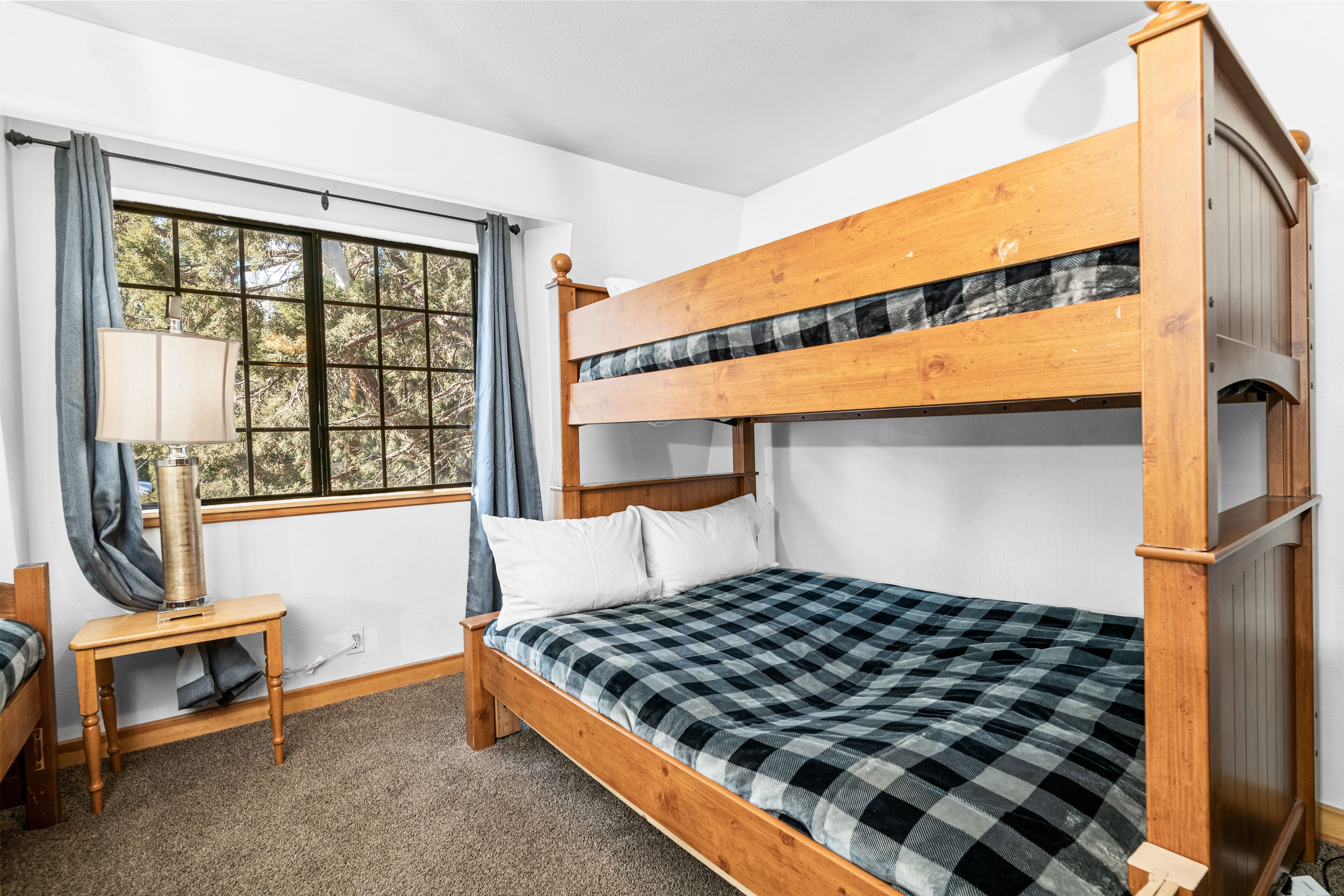 A bunk bed room with a checkered bedspread, a bedside table with a lamp, and a window with curtains.