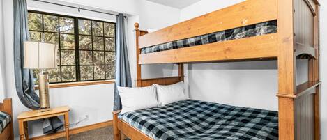 A bunk bed room with a checkered bedspread, a bedside table with a lamp, and a window with curtains.