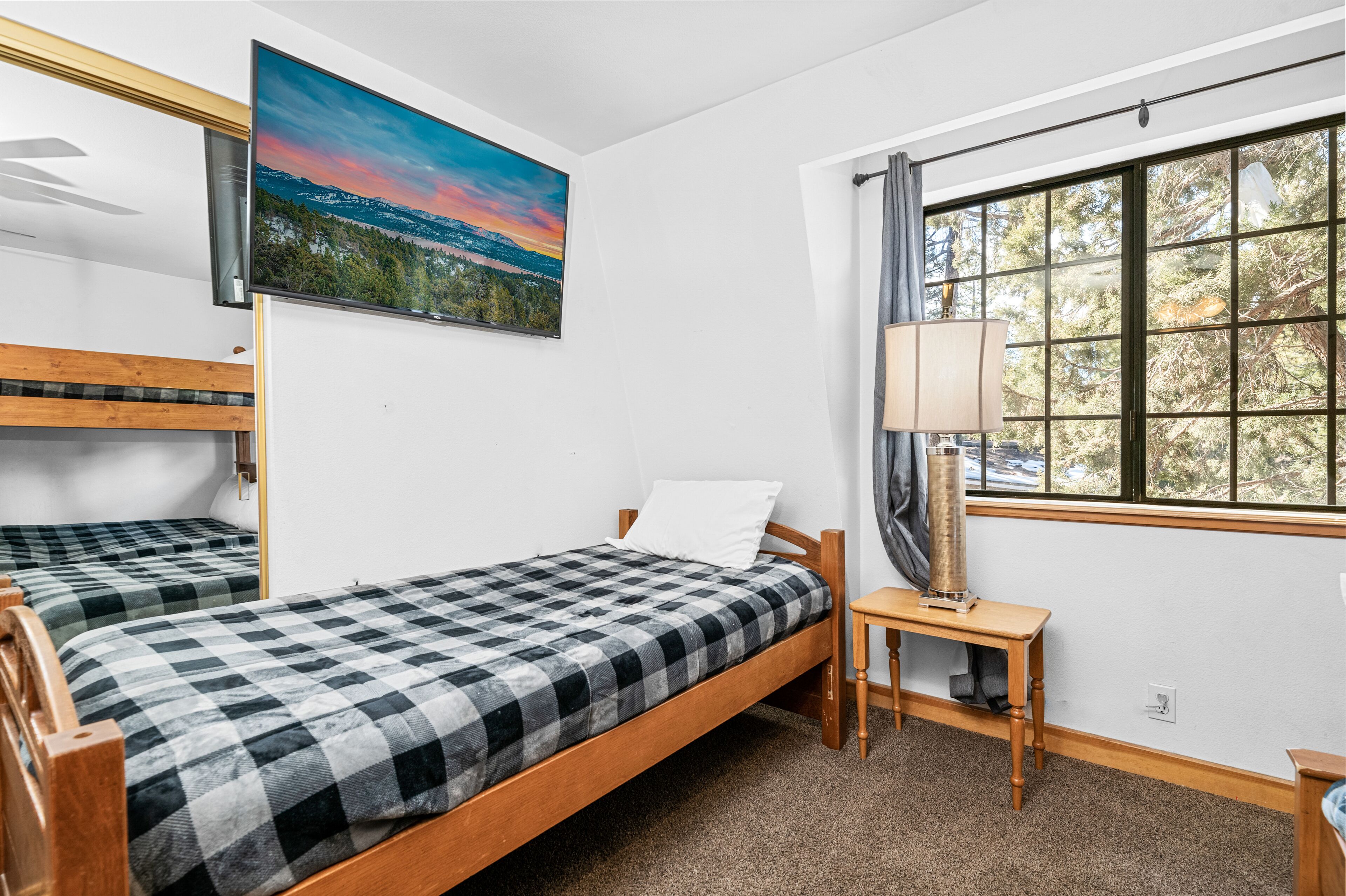 A room with two wooden beds, a painting of a landscape, a window with a view of trees, and a table with a lamp.