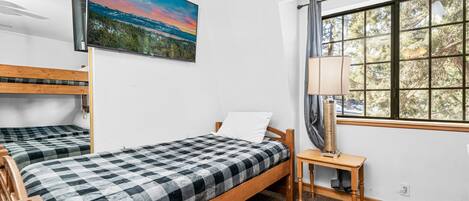 A room with two wooden beds, a painting of a landscape, a window with a view of trees, and a table with a lamp.