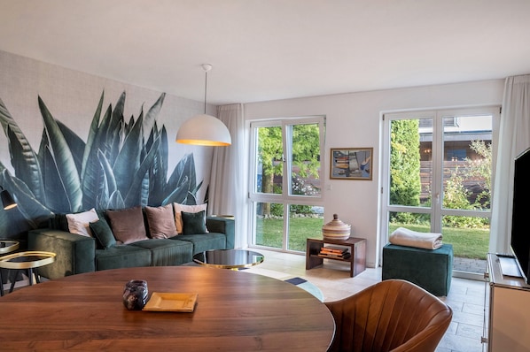 Un salon moderne agrémenté d’une grande fresque murale représentant des plantes, d’une table à manger ronde, d’un canapé vert et de grandes fenêtres offrant une vue sur l’extérieur.
