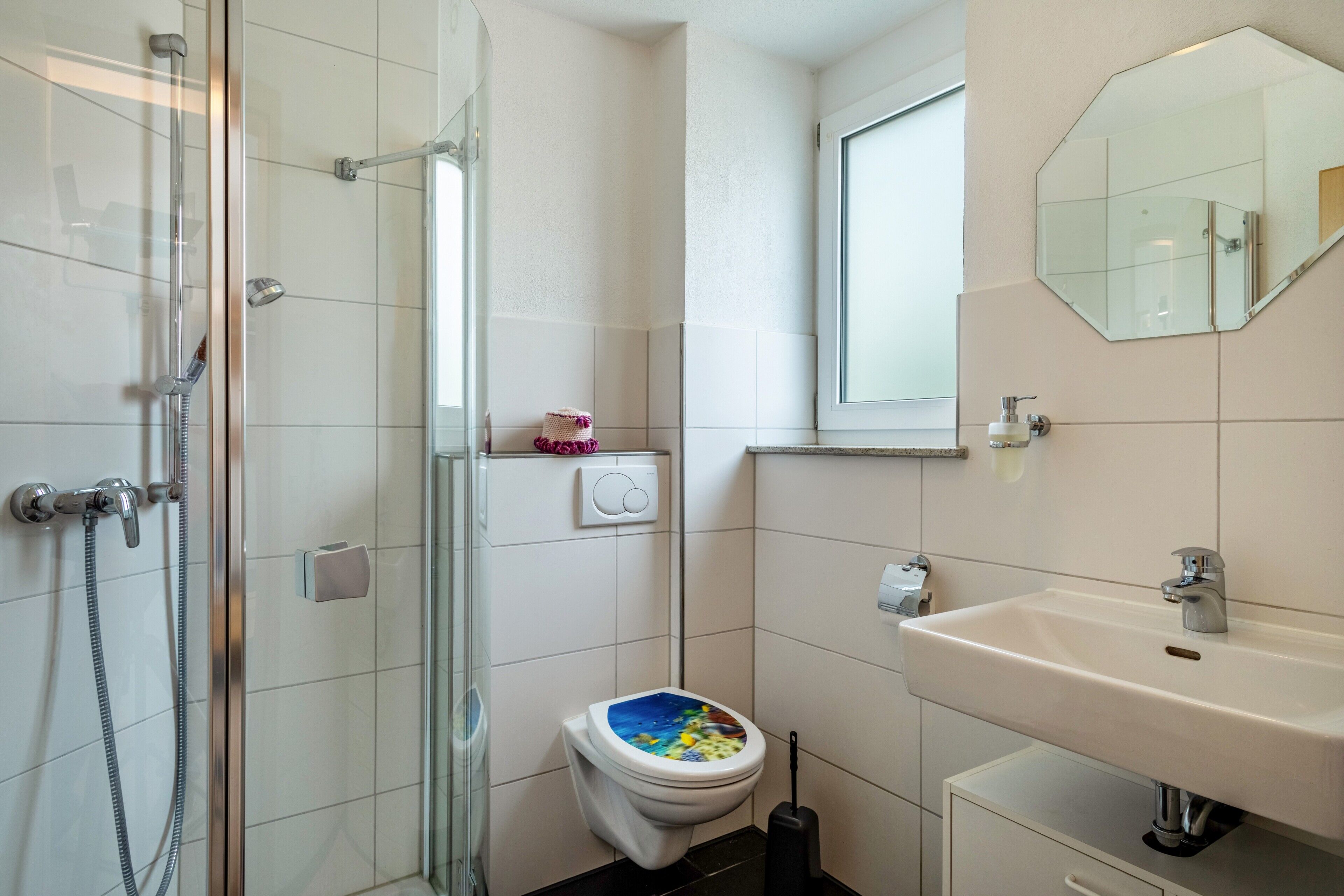 Une salle de bain avec une douche à parois de verre, des toilettes avec un abattant coloré, un lavabo et un miroir.