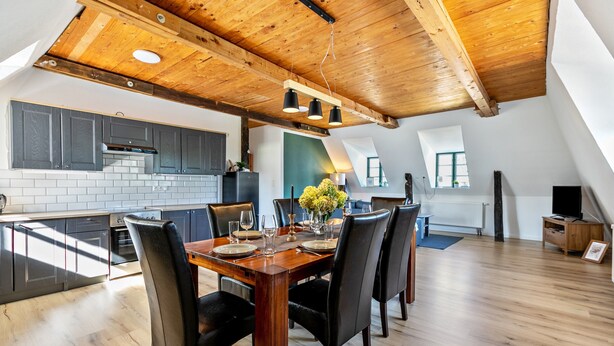 A modern kitchen with dark cabinets, a wooden dining table set for six, and a high ceiling with exposed beams.