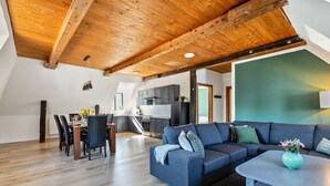 A spacious living area with a wooden ceiling, a dining table with chairs, a sofa, and a kitchen in the background.