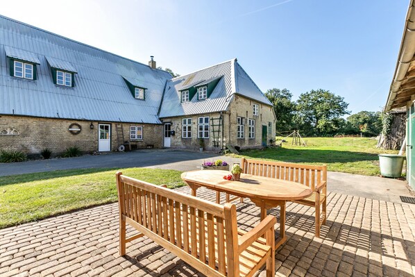 Ein Backsteinhaus mit Metallbedachung, eine gepflasterte Terrasse mit Holztisch und Bank sowie ein Rasenstück.