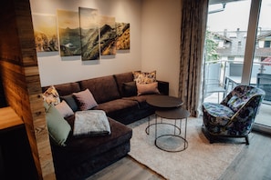 A modern living room with a dark brown sofa, a round coffee table, and a patterned armchair. There are three framed landscape paintings on the wall.