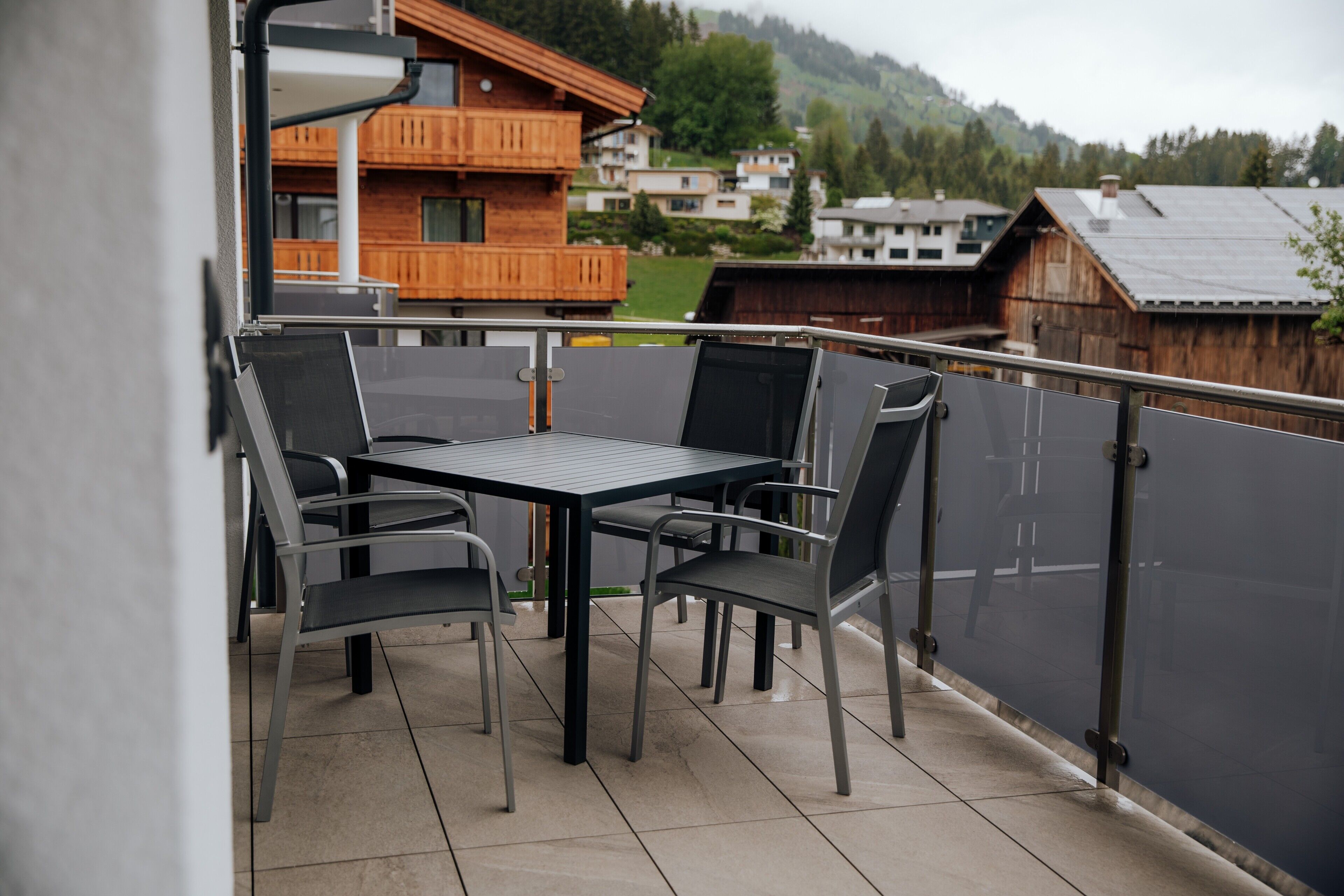 A balcony with a table and chairs, overlooking a residential area with wooden houses and a mountain in the background.